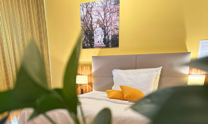 Cozy hotel room with a bed, yellow accents, a plant in the foreground, and a wall art piece.