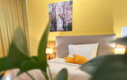 Cozy hotel room with a bed, yellow accents, a plant in the foreground, and a wall art piece.